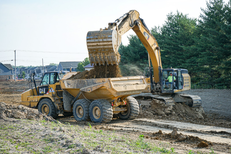 Excavator loading soil into a dump truck at a residential construction site, with another heavy equipment vehicle and houses visible in the background.